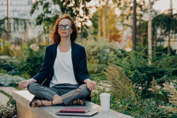 Full length shot of relaxed ginger woman meditates sits in lotus pose wears formal clothes