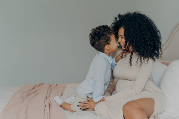 Afro American mother and little son sitting on bed and touching each other with noses