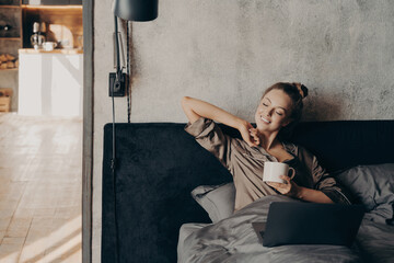 Happy beautiful female with closed eyes drinking coffee in bed before remote work at home - obrazy, fototapety, plakaty