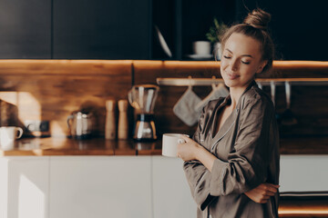 Beautiful young happy woman standing in cozy silk pajamas in kitchen with cup of coffee