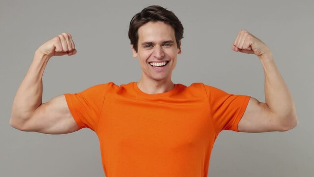 Strong sporty fitness smiling young brunet man 20s years old wears orange t-shirt showing biceps muscles on hand demonstrating strength power isolated on plain grey wall background studio portrait