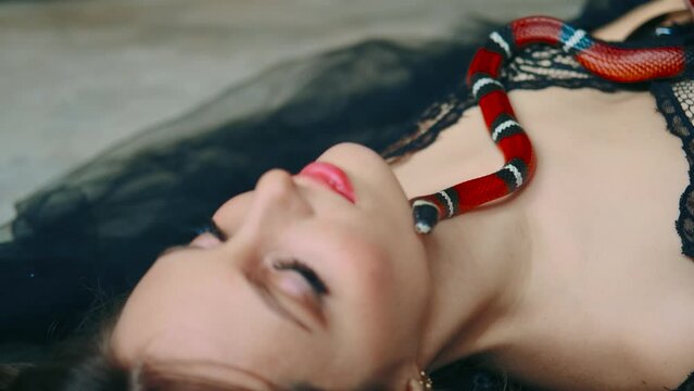 Close-up shot small red snake boa constrictor crawls over body of fantasy woman princess. Sleeping girl, beauty face, eyes closed. Gothic black dress. Lady vampire dark queen in dream. Evening make-up