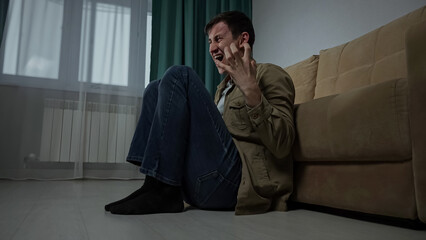 Depressed sad man with hugging the knees sits on floor near sofa in living room. Tired lost guy...