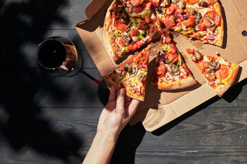 A woman's hand takes a slice of pizza on a sunny day. Pizza in a box craft and a glass of wine. The...