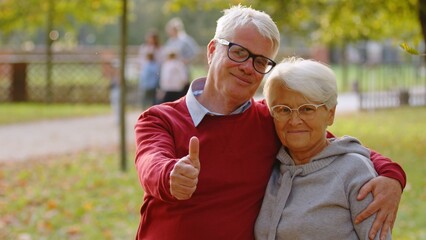 Portrait of aged Caucasian couple in the park both looking at the camera smiling and hugging man showing thumbs up selective focus copy space . High quality photo