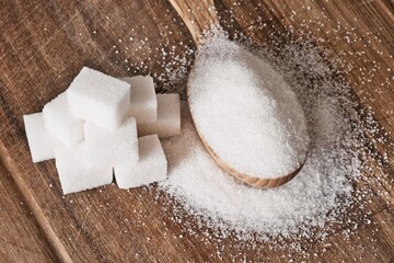 Granulated sugar in wooden spoon and sugar cubes stacked in pyramid on wooden background.