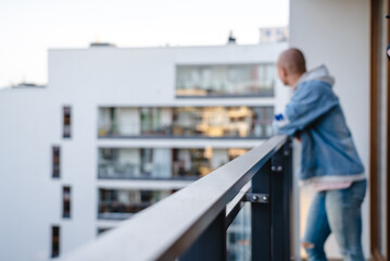 Obraz na płótnie Canvas Blurred photo of a young bald person standing on the balcony, bracing their arms on a guardrail, and looking at the neighborhood. High quality photo