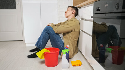 Tired man sits on floor near equipment after cleaning countertop in kitchen at home. Young housekeeper sighs removing rubber gloves from hands