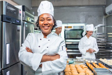 Smiling african  female bakers looking at camera..Chefs  baker in a chef dress and hat, cooking together in kitchen.Team of professional cooks in uniform preparing meals  in the kitchen. - obrazy, fototapety, plakaty