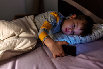 Young boy laying in bed and watching clips by smartphone. Overuse and addiction kids from gadgets. Shot of a young boy using a digital tablet while lying in bed.