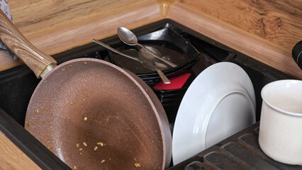 Heap of dirty dishes after cooking breakfast lies in sink waiting to wash. Kitchen utensils with pan and cup on wooden countertop at home closeup
