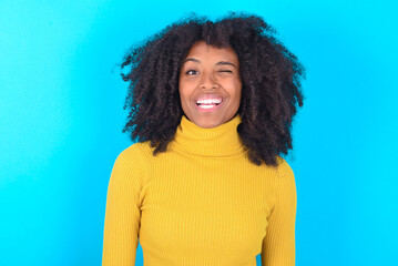 Coquettish Young woman with afro hairstyle wearing yellow turtleneck over blue background  smiling...