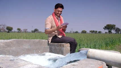 A cheerful Indian farmer counting money while sitting near a tubewell - farm subsidy  bumper crop harvest  agricultural profit  monthly income. Traditional Indian villager doing a thumbs-up gesture...