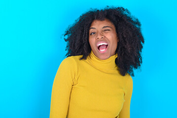 Young woman with afro hairstyle wearing yellow turtleneck over blue background winking looking at...