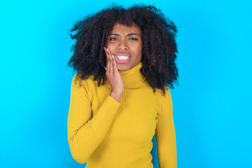 Young woman with afro hairstyle wearing yellow turtleneck over blue background touching mouth with...