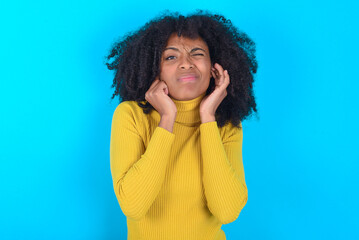 Young woman with afro hairstyle wearing yellow turtleneck over blue background covering ears with fingers with annoyed expression for the noise of loud music. Deaf concept.