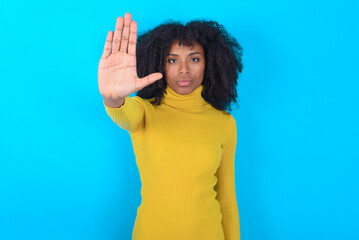 Young woman with afro hairstyle wearing yellow turtleneck over blue background doing stop sing with...