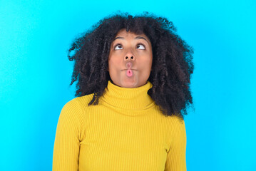 Young woman with afro hairstyle wearing yellow turtleneck over blue background making fish face with lips, crazy and comical gesture. Funny expression.