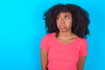 Dissatisfied Young girl with afro hairstyle wearing pink t-shirt over blue background purses lips...