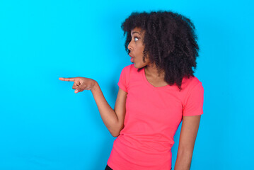 Stunned Young girl with afro hairstyle wearing pink t-shirt over blue background with greatly surprised expression points away on copy space, indicates something