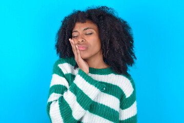 Young woman with afro hairstyle wearing striped sweater over blue background with toothache