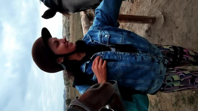 Vertical footage of cheerful young woman in helmet stroking horses on the farm located in the meadow with mountains on background. A smiling lady is caressing mammals. She is going on equestrian walk.