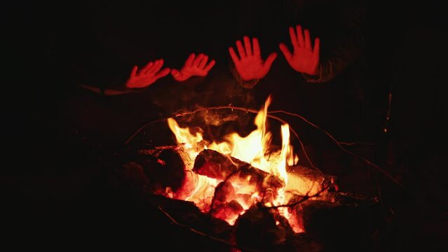 Two people warm their hands by fire on cold night. Frozen tourists, hikers sit near campfire. Backpackers spend time near bonfire. Burning firewood in camping. Campsite, recreation, flame