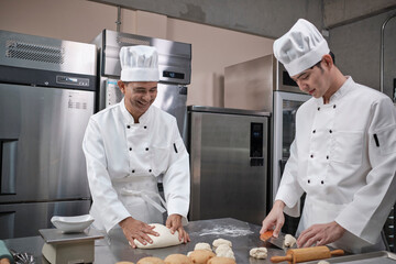 Two professional Asian male chefs in white cook uniforms and aprons are kneading pastry dough and...