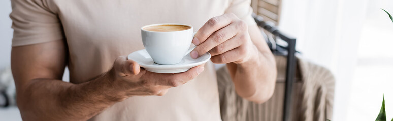 cropped view of man in t-shirt holding saucer with cup of coffee, banner.