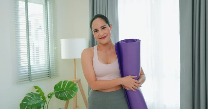 Smiling asian woman holding yoga mat in the bright room.