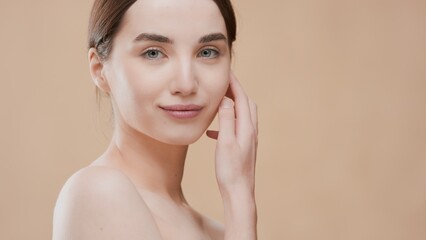 Young slim Caucasian woman with dark hair and naked shoulders touches her jawline face and looks at the camera over her bare shoulder on beige background with water ripples | Skin care concept