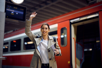 Happy woman waves and greets someone while getting out of train at the station.