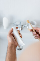 cropped view of man holding toothpaste tube and toothbrush in bathroom.