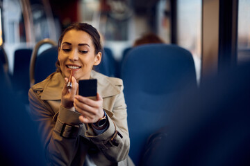 Happy woman uses hand mirror and applies lipstick while commuting by train.