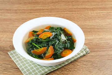 Sayur Bening Bayam, Spinach Clear Vegetable. Indonesian food of spinach, spinach soup with carrots. Served in white bowl on wooden background. 