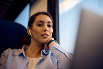 Young woman with earbuds surfing the net on laptop while traveling by train.