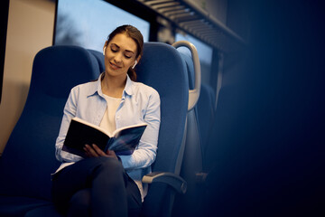 Young smiling woman travelling by train and reading book during the ride.