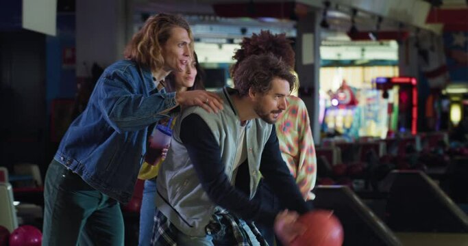 Cinematic shot of happy young male bowler throwing strike ball and exulting victory while having fun to play bowling together with multiracial college friends on weekend in sporting club.