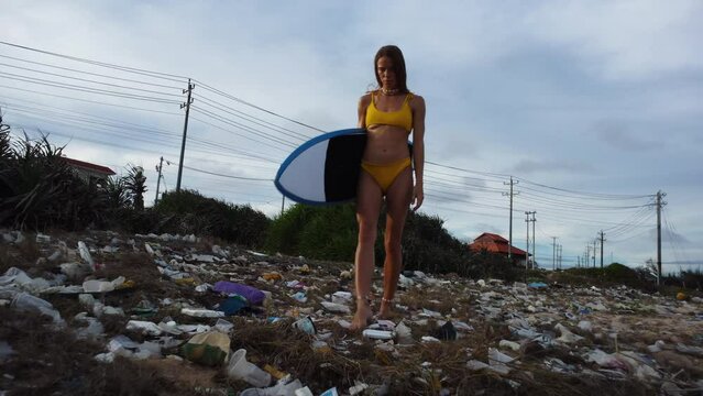 Sexy woman in bikini walk on beach with plastic waste pollution holding surf board, young attractive caucasian surfer in polluted contaminates coast