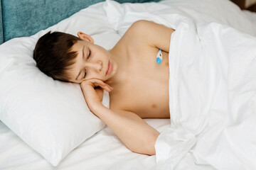 Fototapeta na wymiar A sad sick boy is lying in bed with a thermometer.