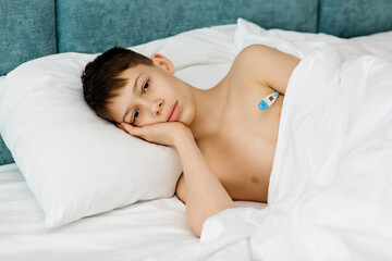 Fototapeta na wymiar A sad sick boy is lying in bed with a thermometer.