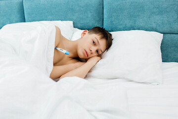 A young man is sick, lying at home under a blanket, holding a thermometer.