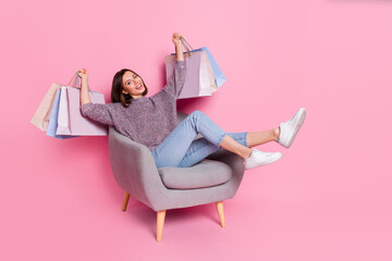 Portrait of attractive cheerful girl sitting in armchair holding bags having fun isolated over pink...