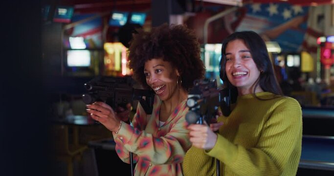 Cinematic shot of young happy interracial girlfriends shot with laser infrared guns while having fun to play shooting simulation game together on fairground in amusement park on weekend.