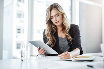 Multitasking makes it happen. Shot of a young businesswoman using a digital tablet and writing notes at her desk in a modern office.