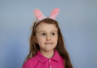 A little girl with bunny ears smiles funny on a blue background. The Easter bunny is a symbol of the Bright Easter holiday. Postcard, copy space