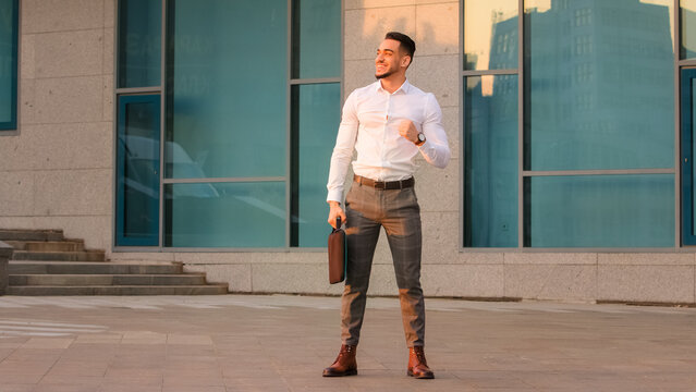 Successful lucky happy arabic spanish business man boss leader candidate entrepreneur with briefcase standing in city near modern building wins achive gets opportunity new job makes victory gesture