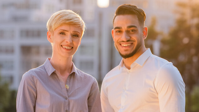 Portrait of multiracial colleagues multiethnic co-workers caucasian business woman blonde short hair and hispanic bearded man stand posing looking at camera smiling in light of sun at sunset sunbeams