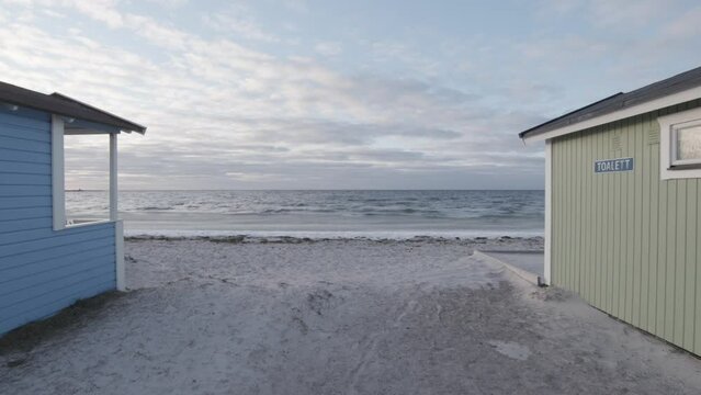 Small cabins by beach cold spring day with iced ocean. Floating gimbal camera forward movement. Eternal sea horizon. White sand and blue sky with clouds. Falsterbo, Sweden. In slow motion high quality