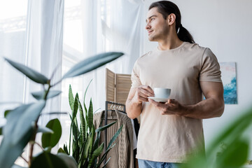 man with long hair holding cup of coffee and saucer near green plants.